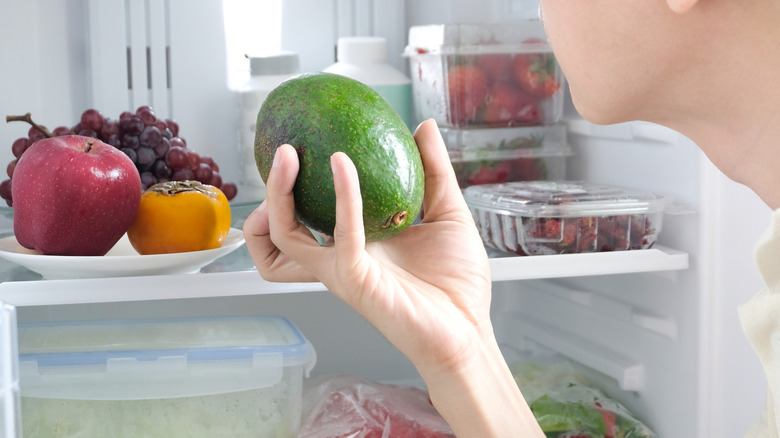A person holds an avocado in front of an open fridge filled with fruit and vegetables.