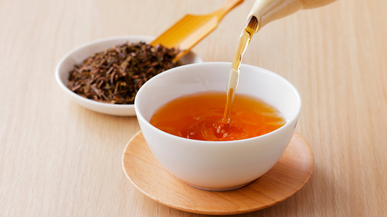 Hojicha tea being poured into a white ceramic cup, with tea leaves being scooped in the background