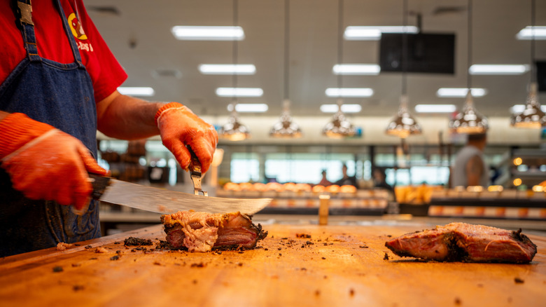 An employee of Buc-ee's slicing smoky & tender brisket on a wooden cutting board.
