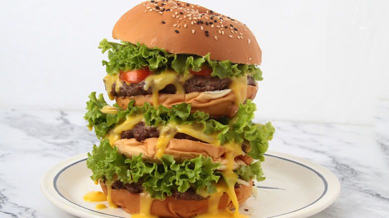 A very tall hamburger on plate with patties, cheese, lettuce, tomatoes