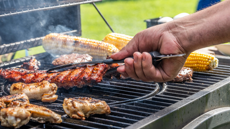 Closeup of a person flipping a rack of ribs on a grill with chicken, corn, and burgers