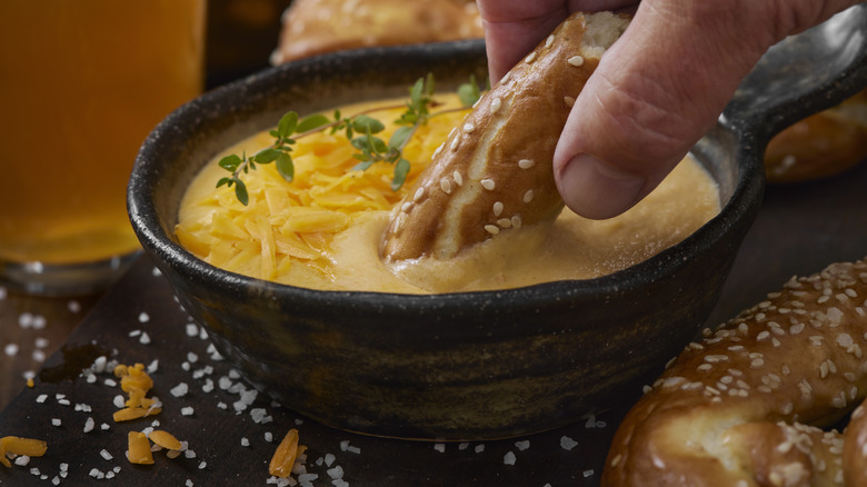 Hand dipping soft pretzel in small bowl of beer cheese dip.