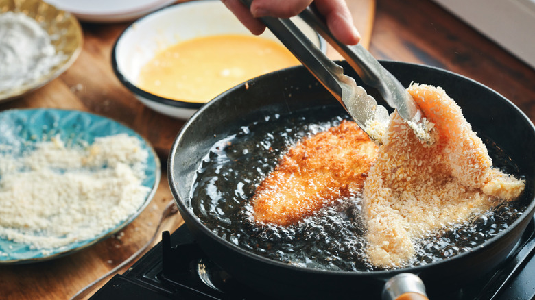 Hand holding frying meat with tongs next to a plate of breading.
