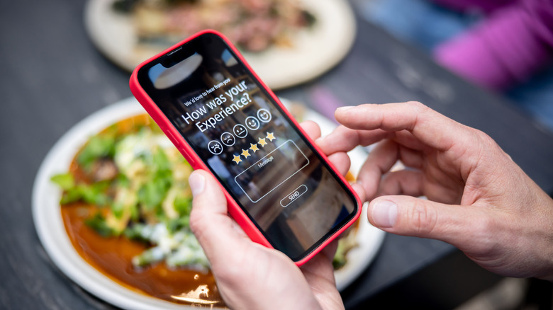 Hands holding red cell phone with customer review interface on screen with plates on table behind