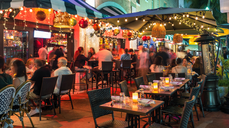 A restaurant patio with string lights and umbrellas at night with a stucco building and palm trees behind