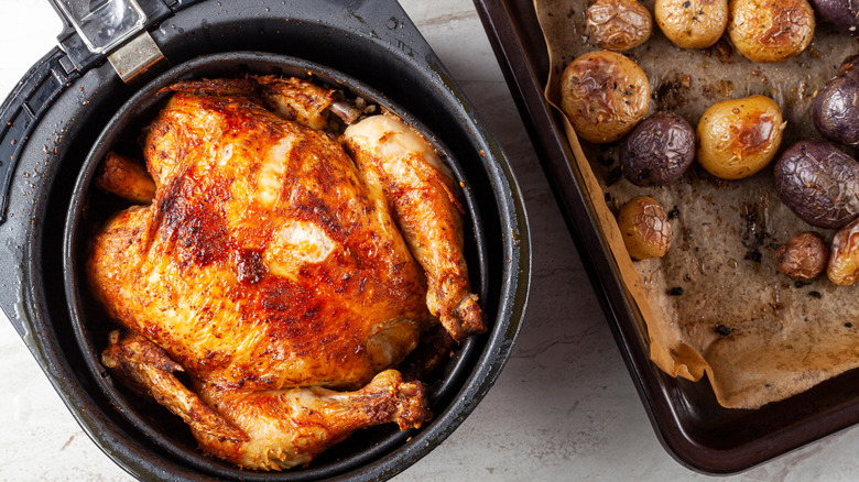 Two air fryer baskets, one with a chicken roasting inside, and another with a liner and potatoes on it