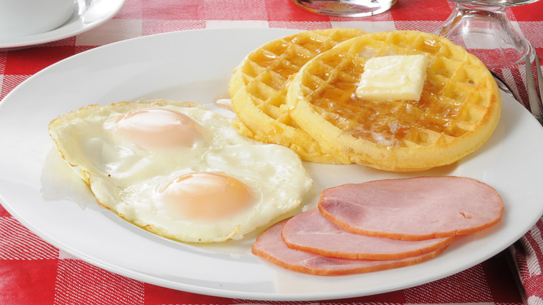 A white plate with two fried eggs, two waffles, and three slices of Canadian bacon