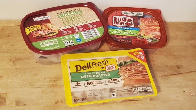 three packages of sliced turkey meat on wooden surface, including brands like Lunch Mate, Hillshire Farms, and Oscar Mayer