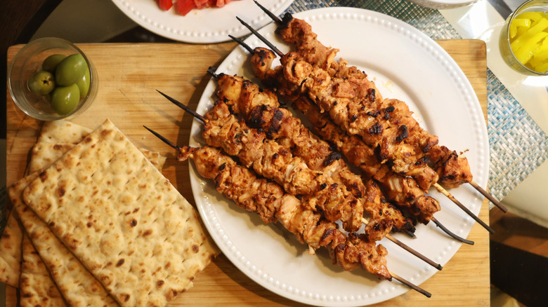 overhead view of plate of chicken kebabs on cutting board next to flatbreads and a bowl of green olives