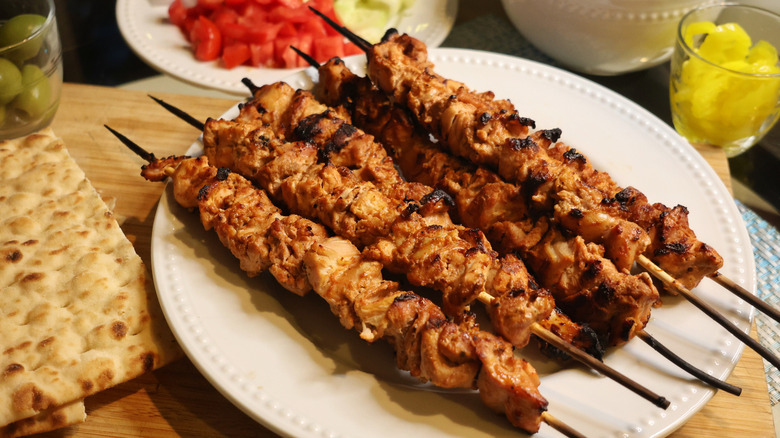 chicken kebabs on a plate surrounded by flatbreads, olives, and vegetables