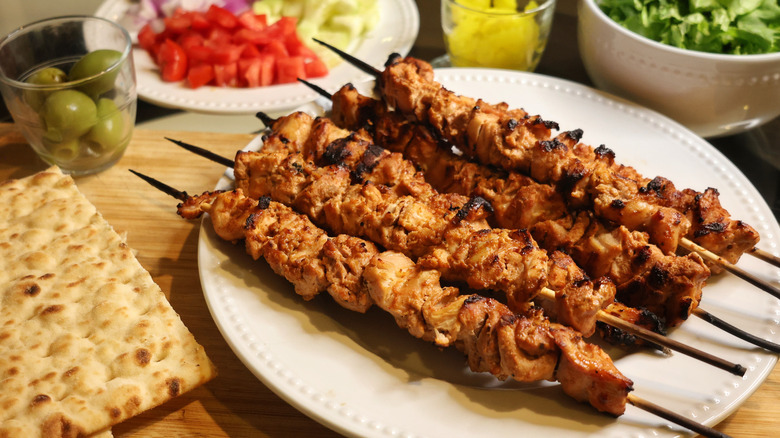 plate of chicken kebabs on cutting board next to flatbreads, olives, and vegetables