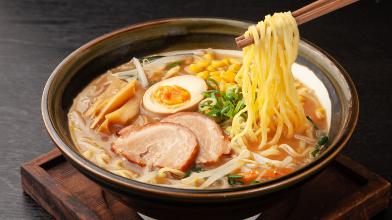 Chopsticks taking noodles out of a heaping bowl of ramen noodles with meat, corn, green onions, and a hard boiled egg