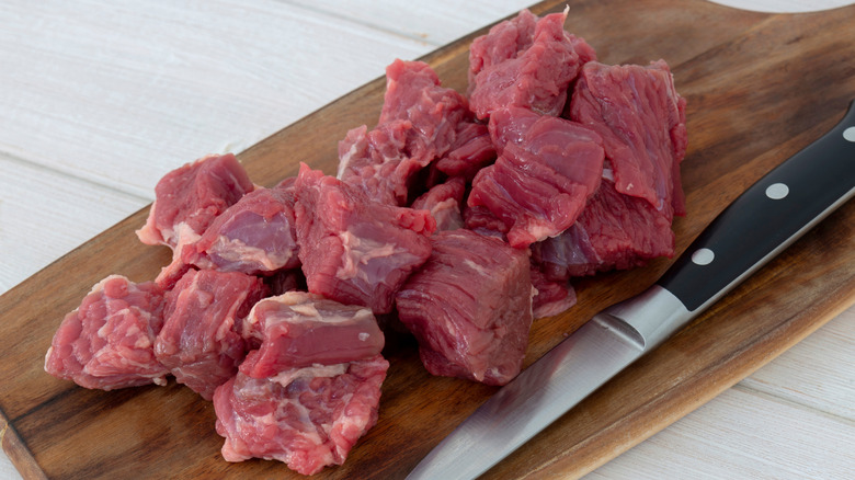 Fresh, pink, raw beef brisket cut into cubes on top of a wooden cutting board with a knife to the side.