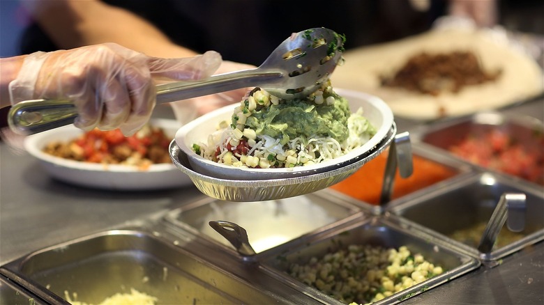 Gloved hand holds a Chipotle burrito bowl and scoops toppings onto the top