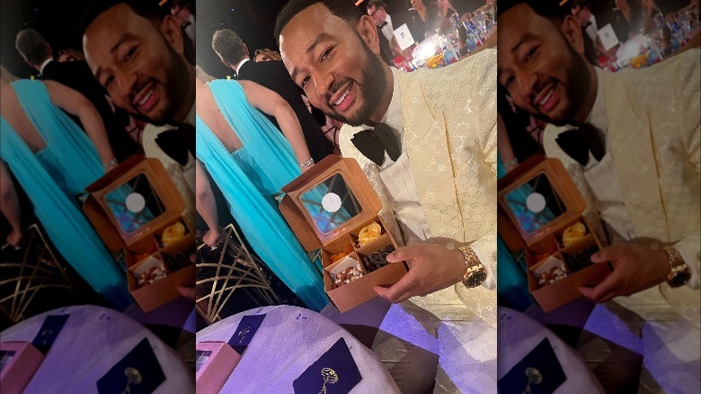 john legend in white suit and black bow tie at emmy awards, he's holding a box of nuts, dried fruit, chocolate-covered pretzels