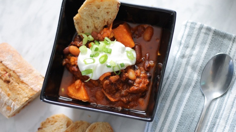 square bowl of chili with sour cream, baguette slices and green onions