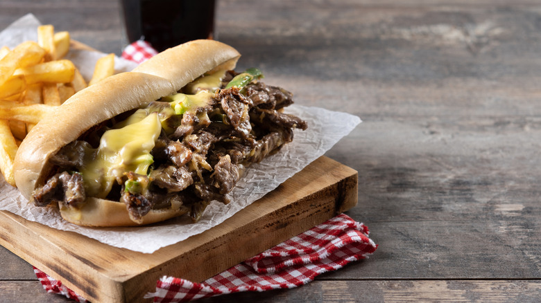 A classic Philly cheesesteak on a hoagie roll with melted cheese served on a piece of parchment paper on a wooden board, with french fries and a dark drink in the background
