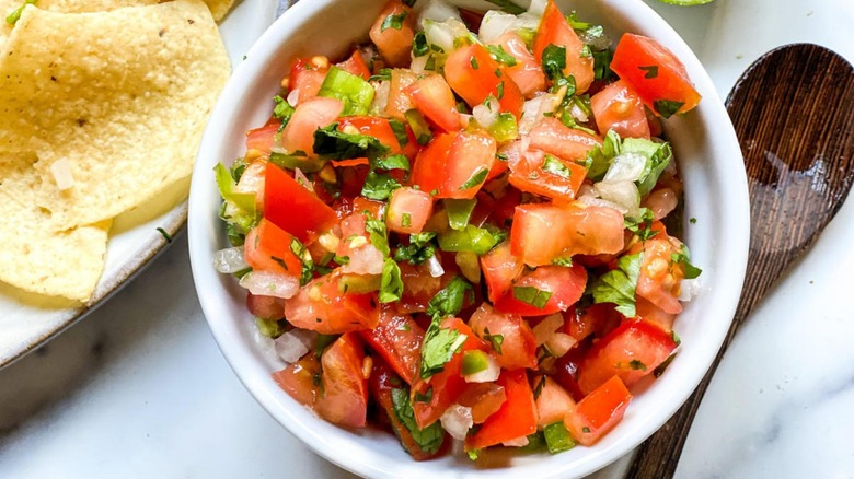 Overhead shot of a white bowl filled with pico de gallo next to a wooden spoon and a tomato