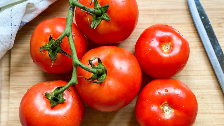 Overhead shot of six red tomatoes (four with the stems still on) next to a knife on a wooden cutting board