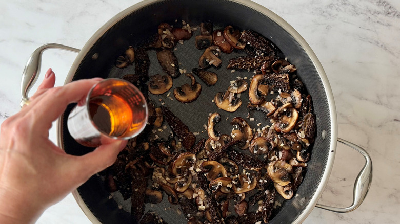 Hand holding small glass of cognac over skillet with mushrooms