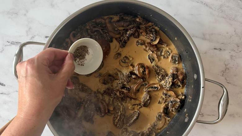Hand holding small bowl of salt and pepper over creamy mushroom sauce in skillet