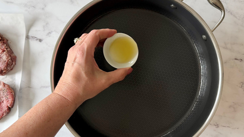 Hand holding small bowl of oil over skillet