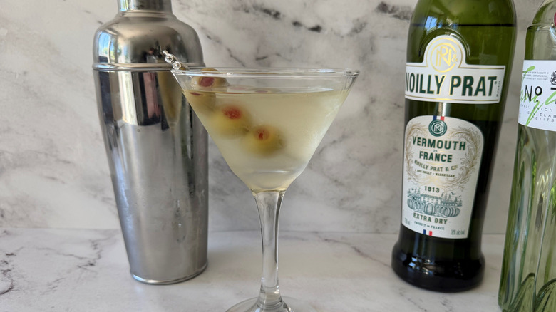 Martini with olive garnish next to cocktail shaker and liquor bottles