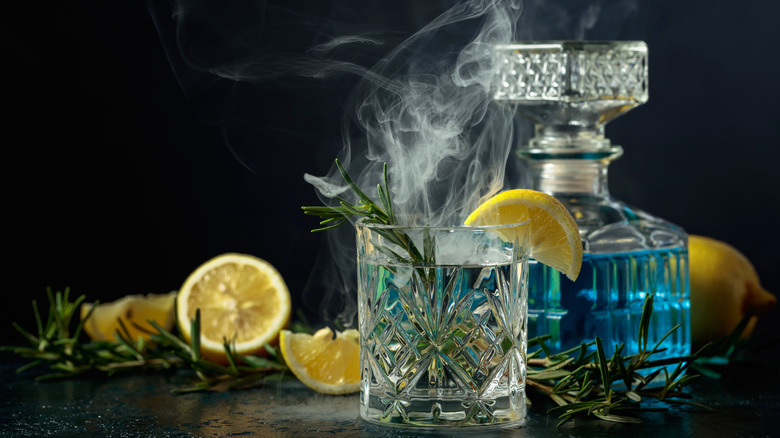 A glass of clear spirit, infused with smoked rosemary and lemons in front of a decanter