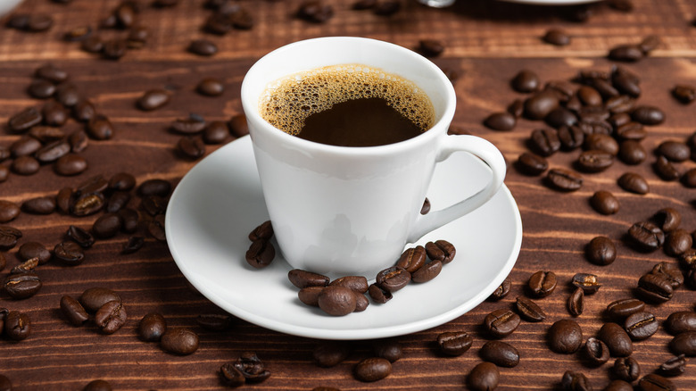 White cup of coffee on white saucer surrounded by coffee beans.