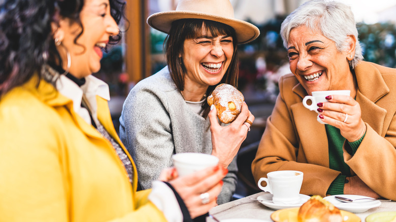 Three adults laughing while two hold coffee cups and one holds a pastry.