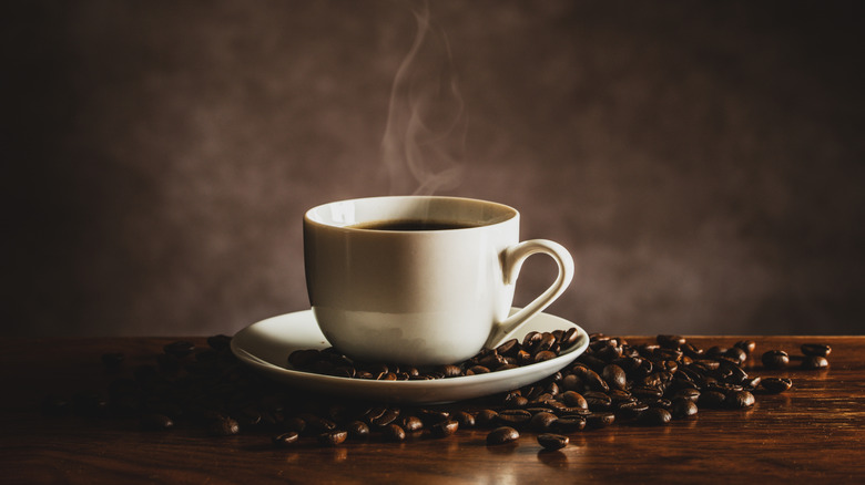 White cup of steaming-hot coffee on saucer plate with beans on wooden table against gray-brown wall.
