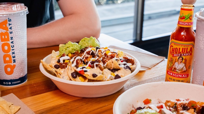 Qdoba nacho bowl with guacamole, corn, and beans, on a table next to a soft drink and hot sauce