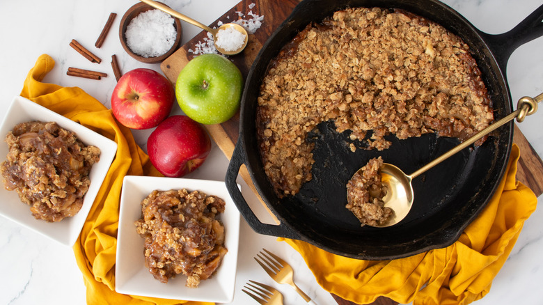 cooked apples with crumbly brown topping in two square white bowls next to a black frying pan partially full of baked oat mixture