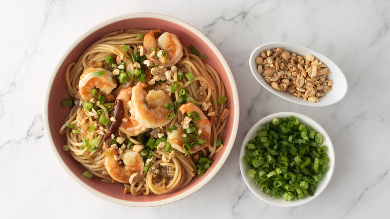 Kung pao spaghetti with shrimp in bowl next to small bowls of peanuts and scallions