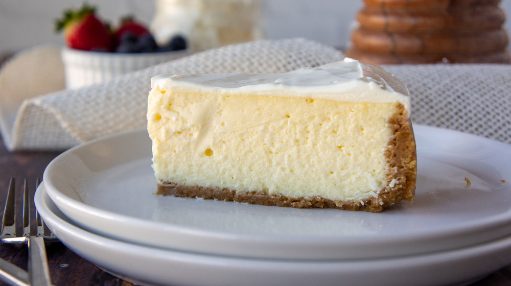 slice of vanilla cheesecake on a white plate in front of a bowl of berries and whipped cream