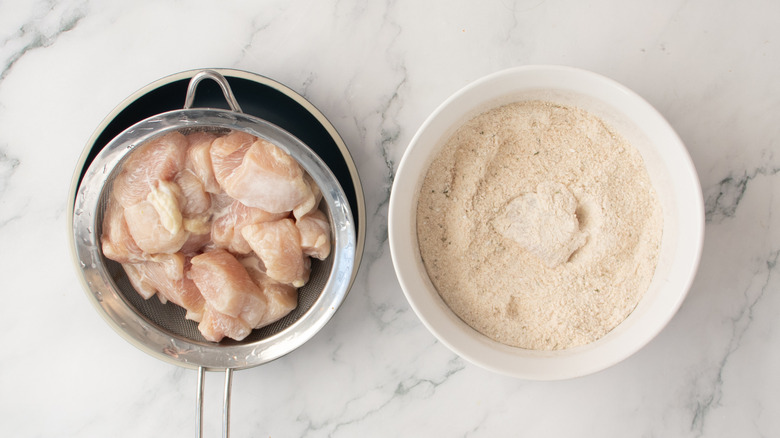raw chicken pieces on a colander next to a bowl of seasoned flour
