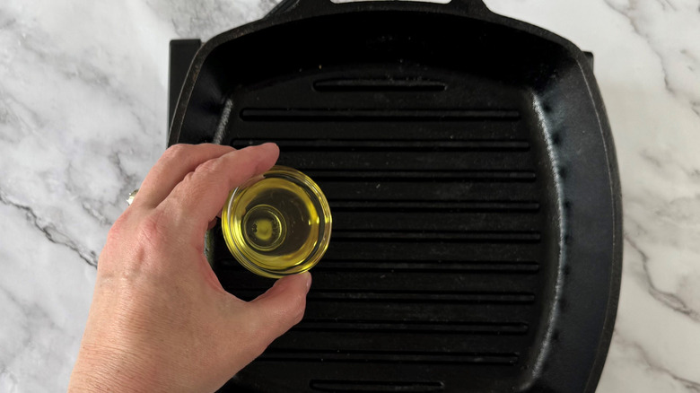 Hand pouring small bowl of oil onto grill pan