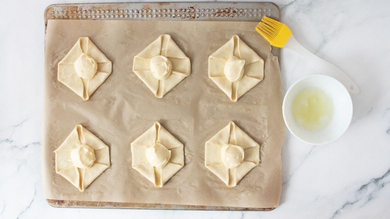 Unbaked danishes on baking sheet next to pastry brush and egg white in bowl
