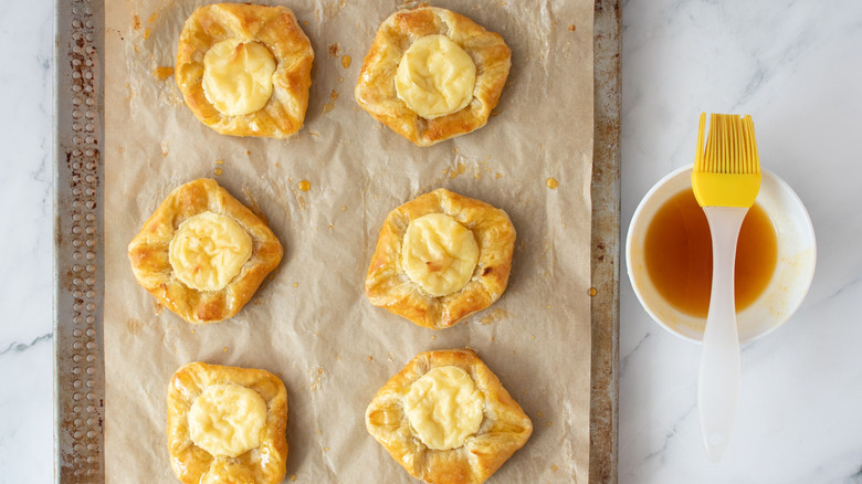 Cream cheese danishes on baking sheet next to apricot jelly in bowl with pastry brush on top