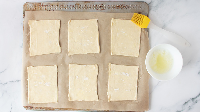 Puff pastry squares on baking sheet next to pastry brush and egg white in bowl