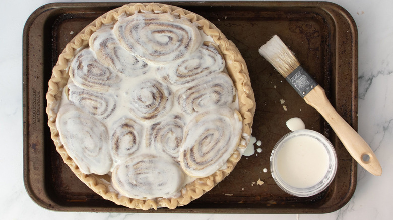 Cinnamon rolls topped with cream in pie crust next to pastry brush and bowl of cream