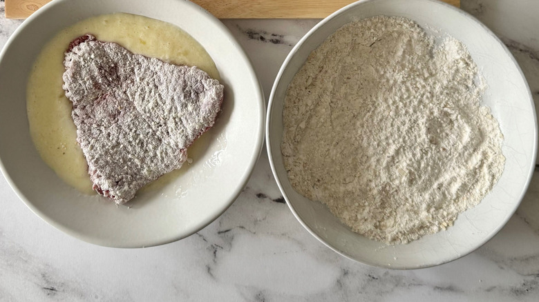 Flour-coated steak in bowl with egg mixture next to bowl with flour mixture