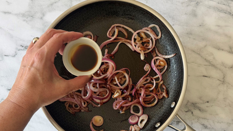 Hand holding small bowl of Worcestershire sauce over grilled onions in pan