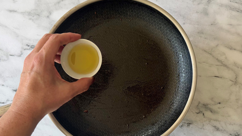 Hand holding small bowl of oil over skillet