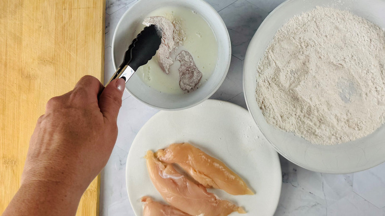 Chicken dredging station with tongs dunking tender into buttermilk