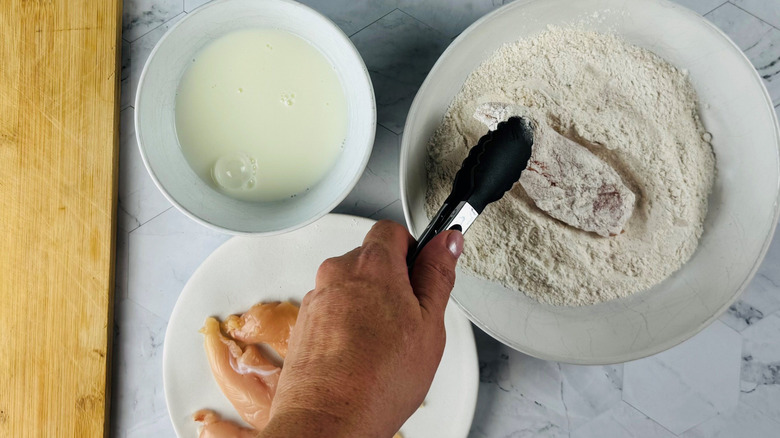 Chicken dredging station with tongs dipping tender into flour