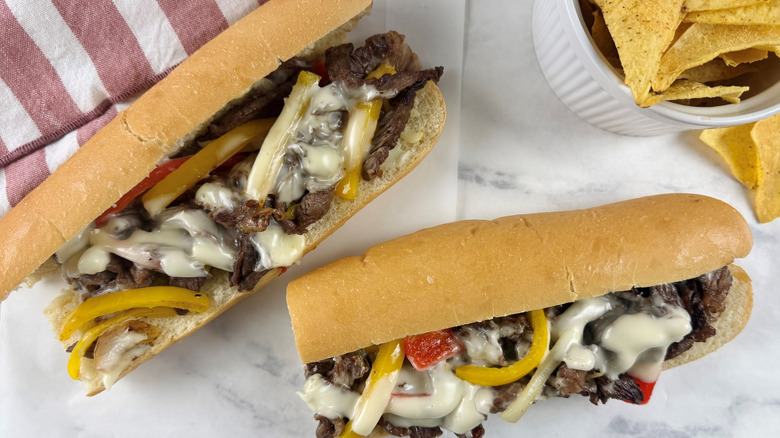 steak and cheese sandwiches with bell peppers on a white countertop next to a striped napkin