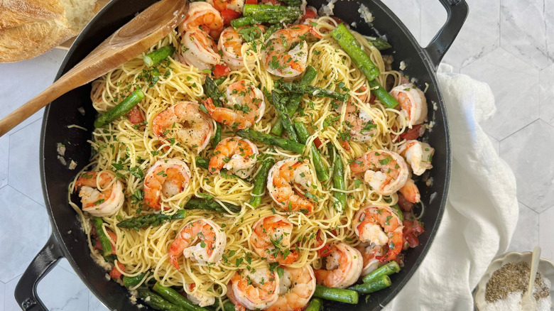 Shrimp scampi with asparagus and pasta in large pan next to bread and seasoning