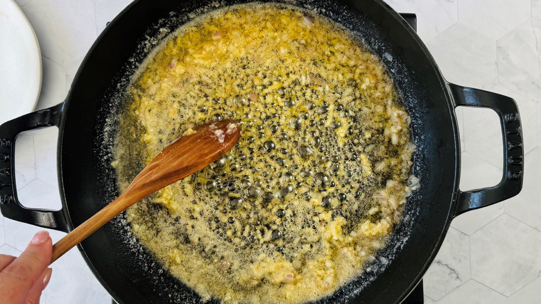 Hand using wooden spoon to stir garlic and shallots in melted butter in pan