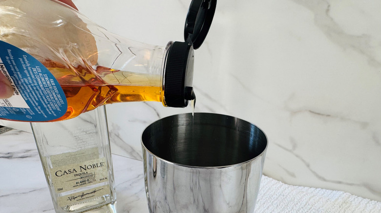 hand pouring brown liquid from a bottle into a cocktail shaker